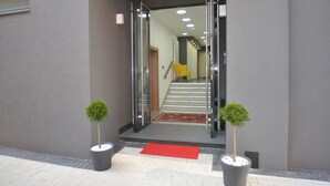 A modern building entrance with a glass door, a red carpet, and two potted plants. - Hotel Family (Sarajevo)