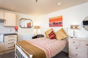 Quarto com cama, mesas de cabeceira, espelho, pintura e área de cozinha com armários e eletrodomésticos.