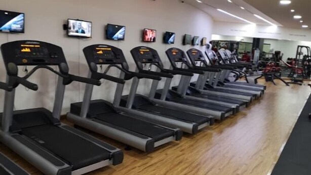 Une rangée de tapis de course dans une salle de sport, avec des appareils d’exercice et des téléviseurs accrochés au mur.