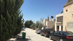 Une rue résidentielle bordée de voitures garées, avec une poubelle verte et des immeubles dotés d’antennes paraboliques.