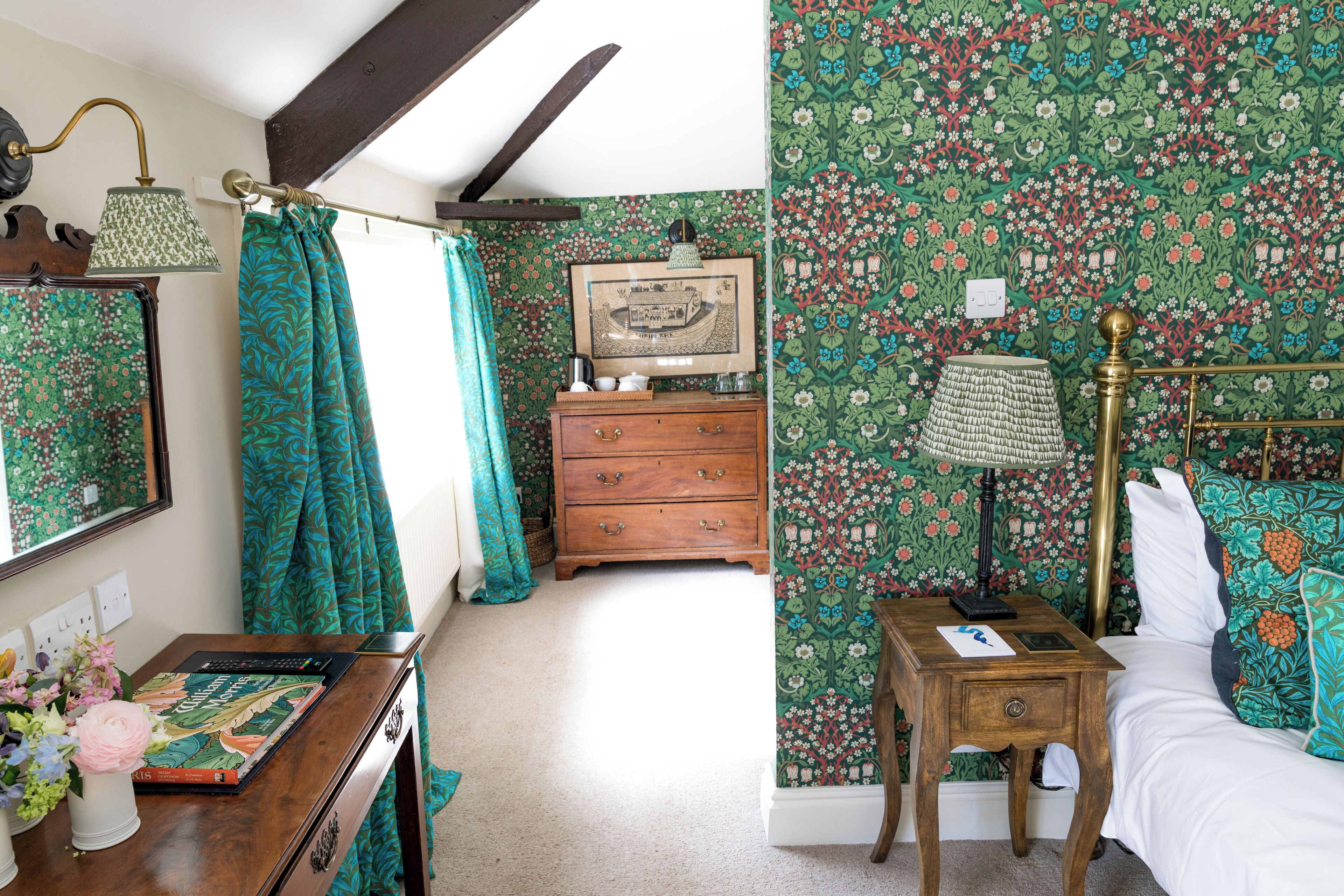 Une chambre à coucher avec un papier peint à motifs floraux, une commode en bois, une table de chevet avec une lampe et un lit agrémenté d’oreillers à motifs.