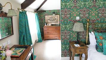 Une chambre à coucher avec un papier peint à motifs floraux, une commode en bois, une table de chevet avec une lampe et un lit agrémenté d’oreillers à motifs.