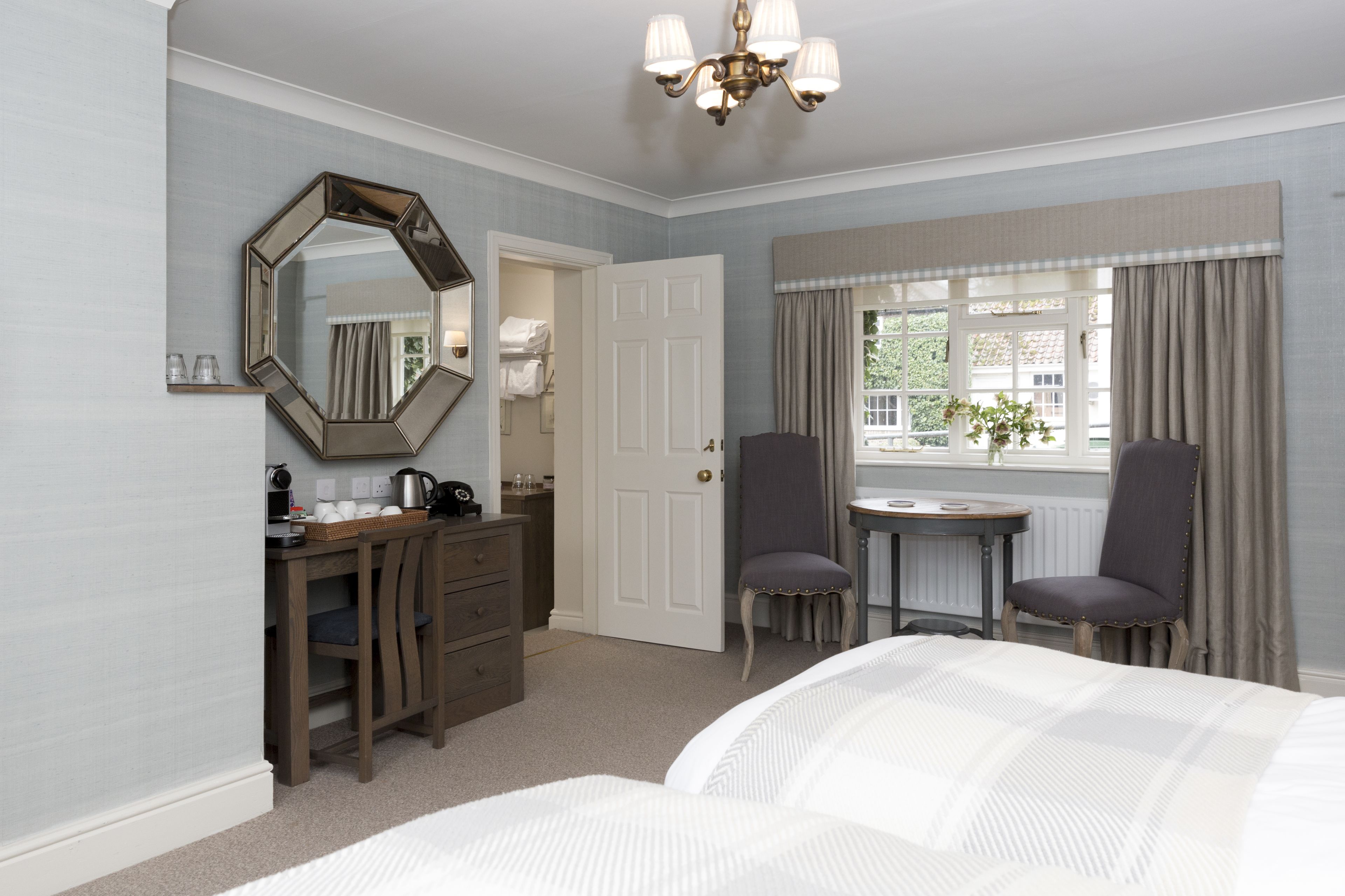 A bedroom with a bed, a desk, a chair, a mirror, a window with curtains, and a chandelier.