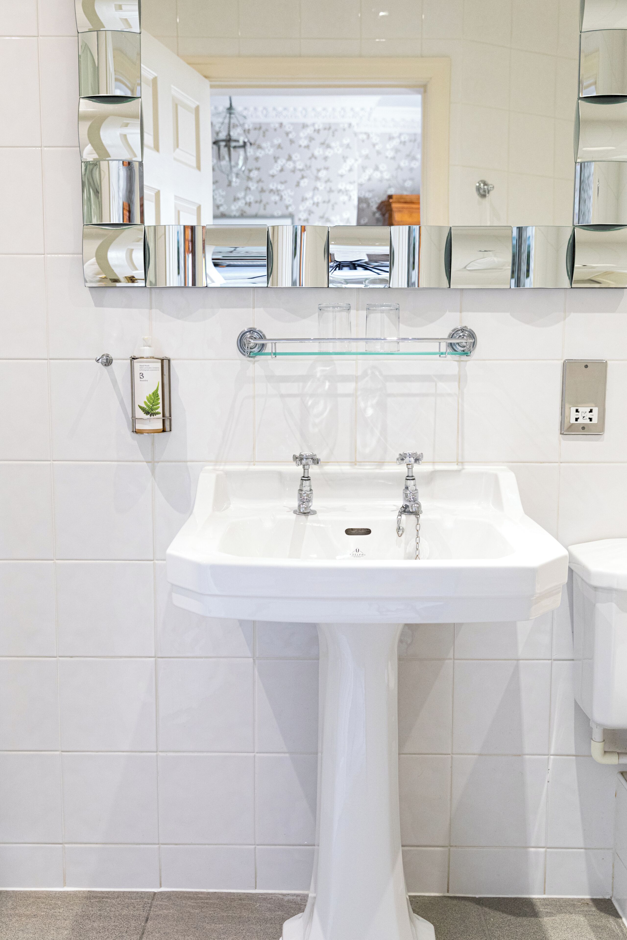 Un baño con un lavamanos blanco de pedestal, un estante de vidrio sobre el lavamanos y un espejo que refleja la habitación.