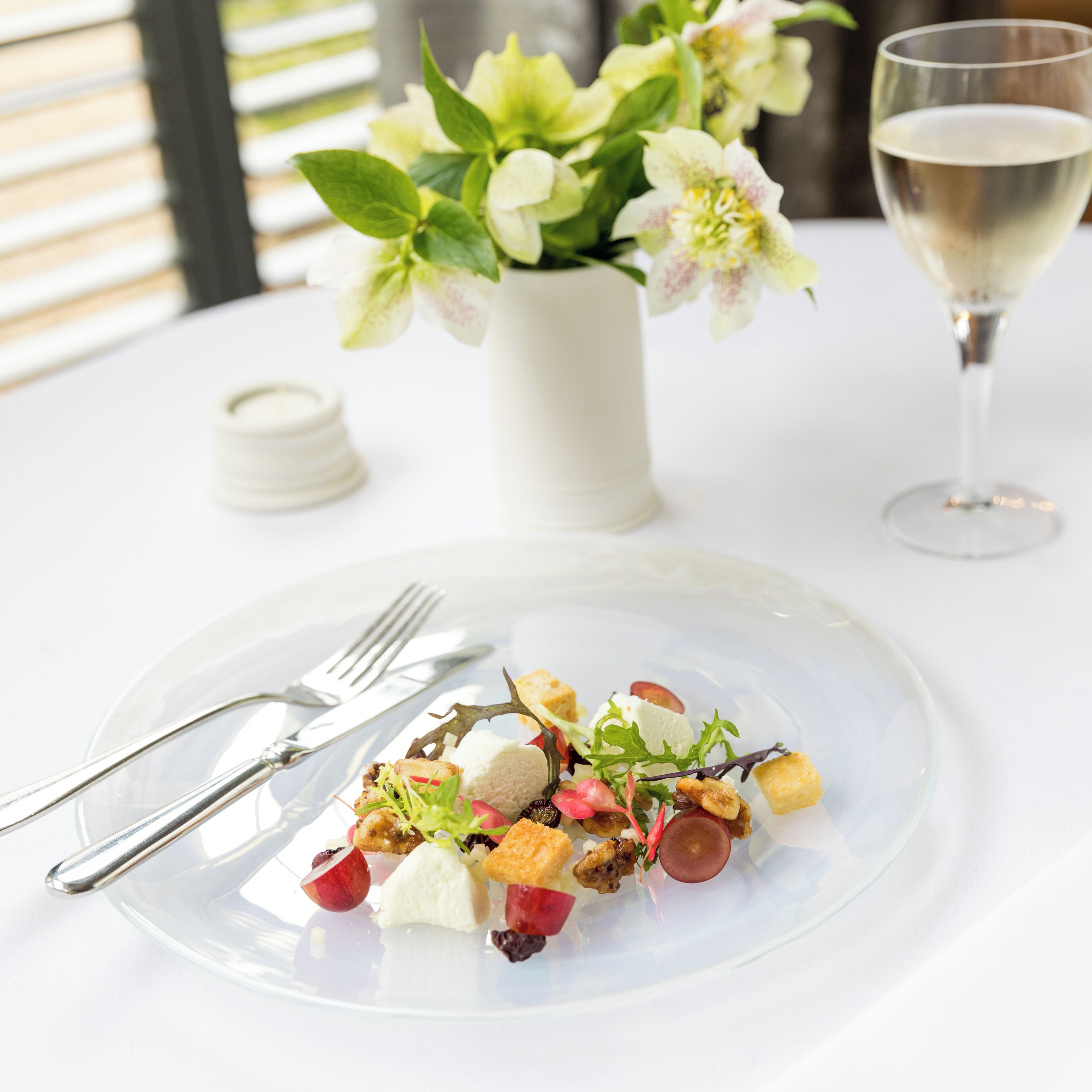 A dining table with a clear glass plate of food, a white vase with flowers, and a glass of white wine.