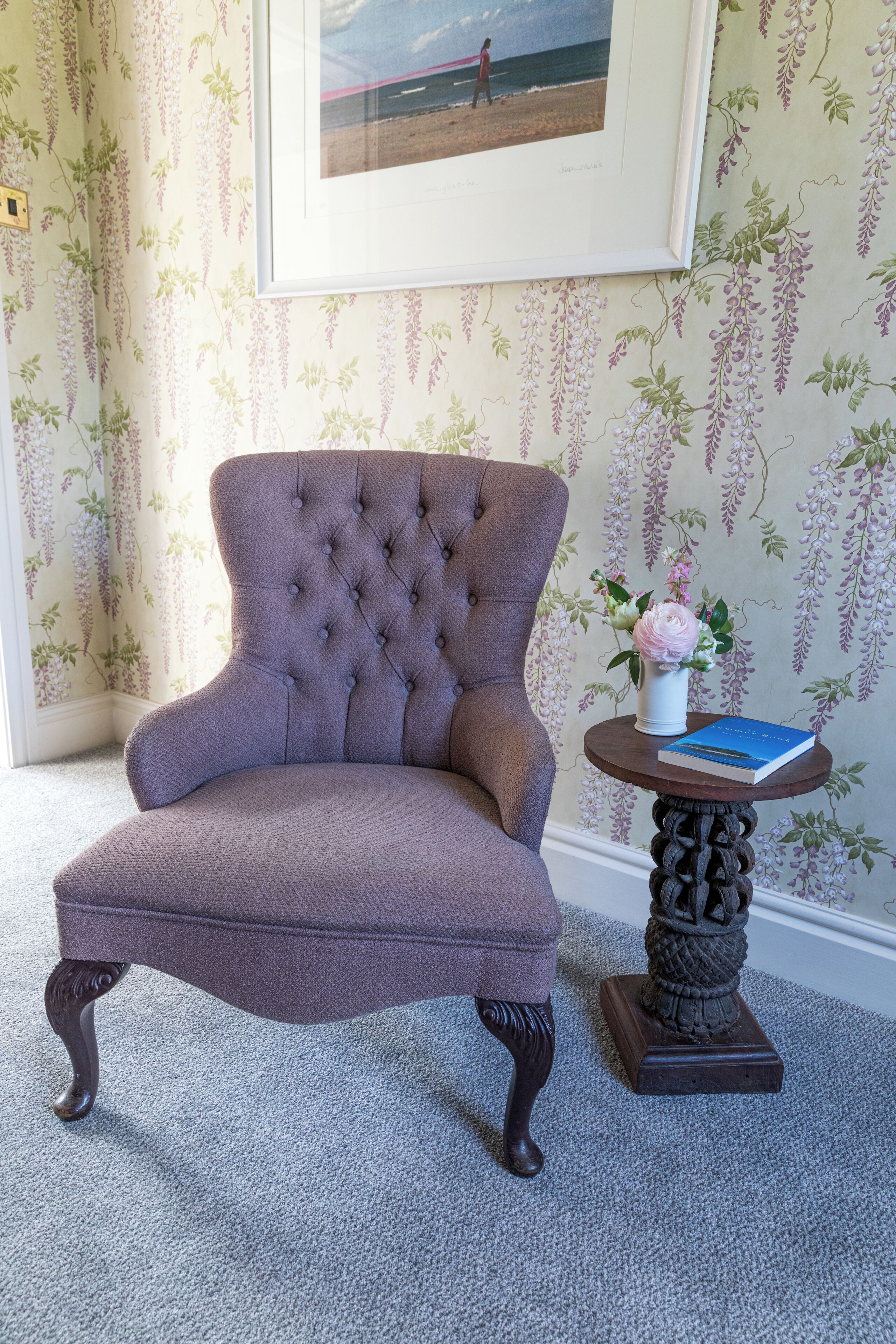 A tufted armchair with a dark fabric, a small round table with a vase and book, and wallpaper with hanging floral patterns.