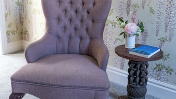A tufted armchair with a dark fabric, a small round table with a vase and book, and wallpaper with hanging floral patterns.