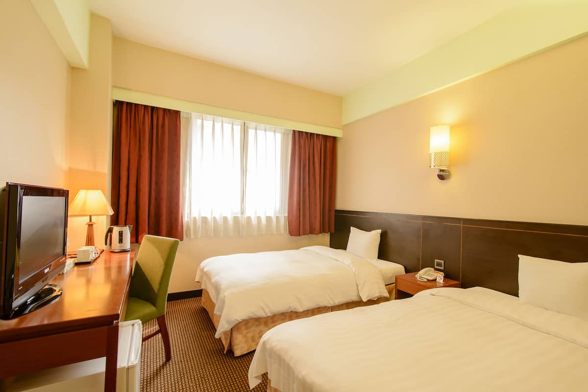 A hotel room with two beds, a desk with a computer and lamp, a chair, and a window with curtains.