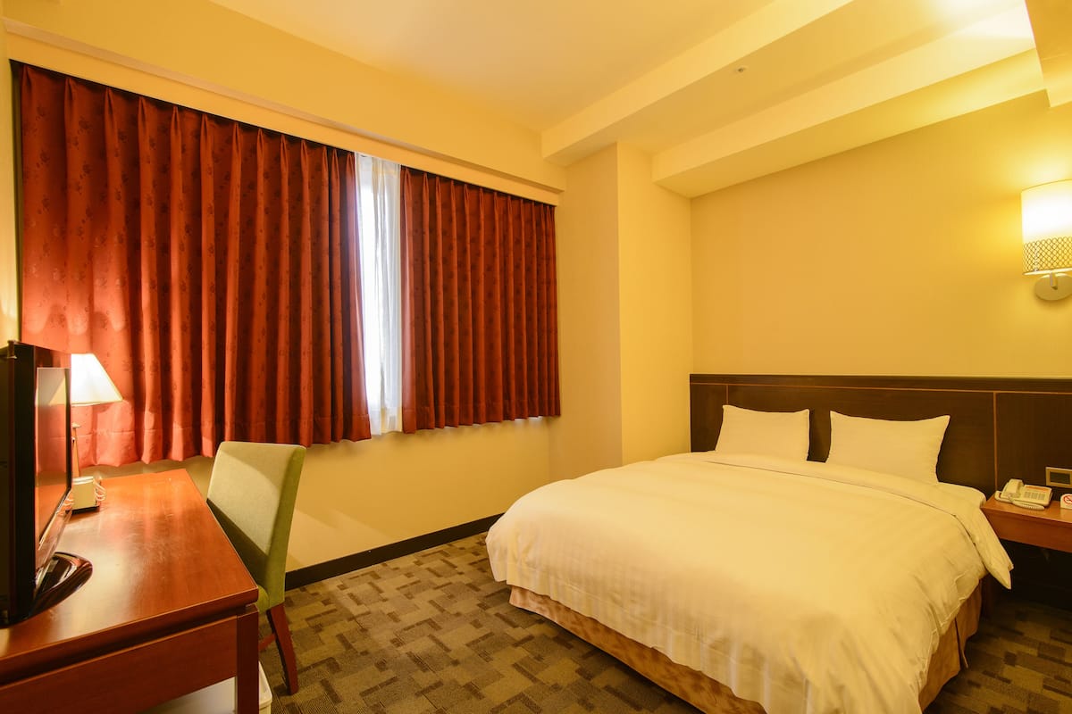 A hotel room with a bed, a desk, a chair, a lamp, and a telephone.