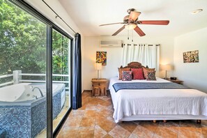 Une chambre avec une grande fenêtre, un ventilateur de plafond, un lit agrémenté d’oreillers à motifs et une petite table de chevet avec une lampe.