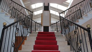 Una imponente escalera con peldaños alfombrados de rojo, barandillas de hierro forjado negro y un tragaluz en la parte superior.