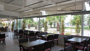 Amplio espacio interior con mesas y sillas dispuestas para comer, ventanas grandes que ofrecen vistas a palmeras y un estacionamiento, y un techo con vigas y luminarias a la vista.