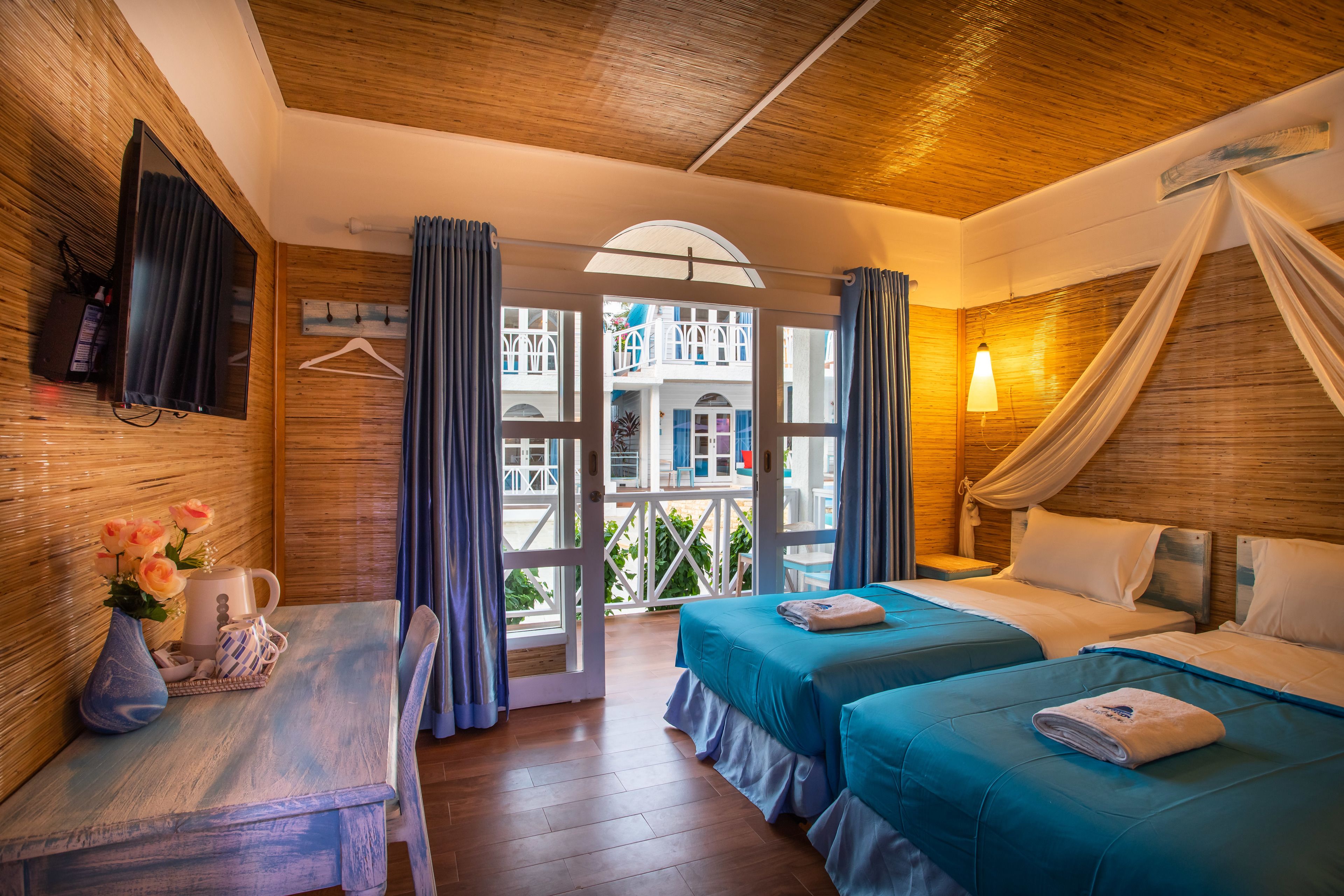 A hotel room with two beds, a wooden ceiling, a TV, a table with a vase of flowers, and a view of a building outside.