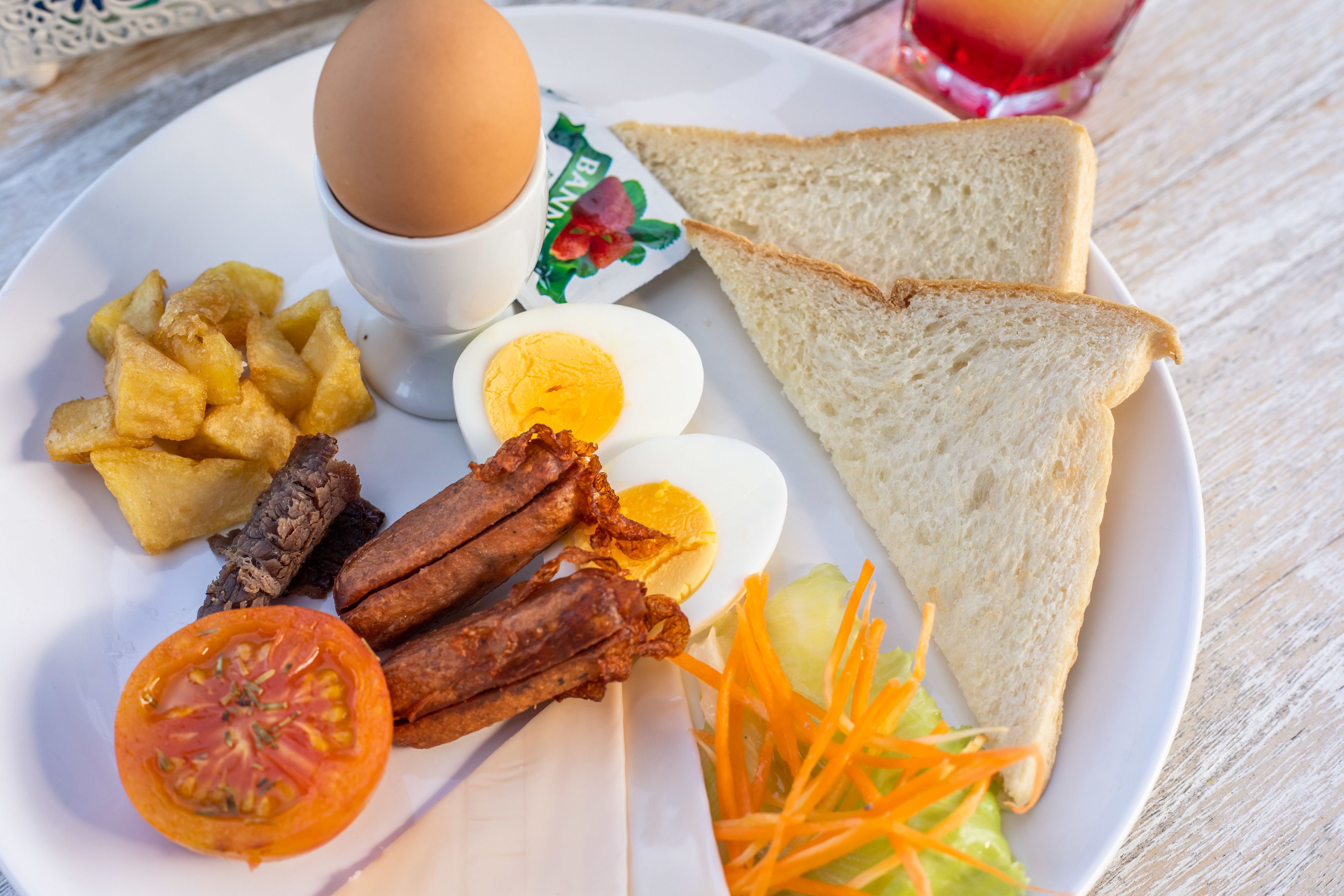 A plate with a boiled egg, bacon, fried potatoes, tomato slice, shredded carrots, and two slices of white bread.