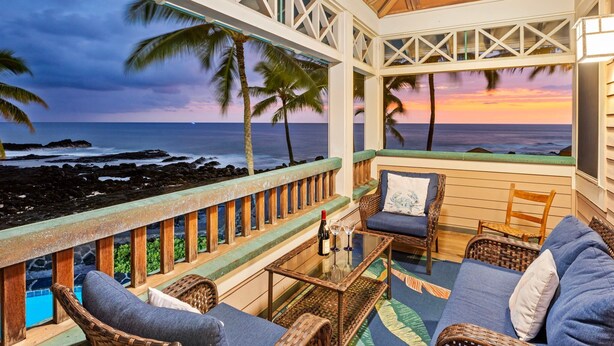 A beachside balcony with wicker furniture, a glass table, and a view of the ocean at sunset. - 🏖️Gorgeous Ocean Front Estate | Gated Community | Walk to Town (Kailua Kona)