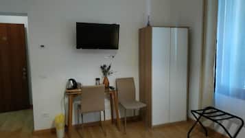 Ett litet rum med trägolv, ett skrivbord med en stol, en tv som är monterad på väggen, en garderob och ett fönster med gardiner.