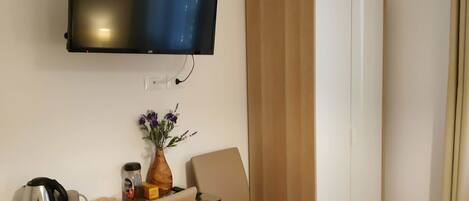 Ett hotellrum med ett skrivbord i trä, två stolar, en tv som är monterad på väggen, en garderob och ett litet bord med en vas med blommor.