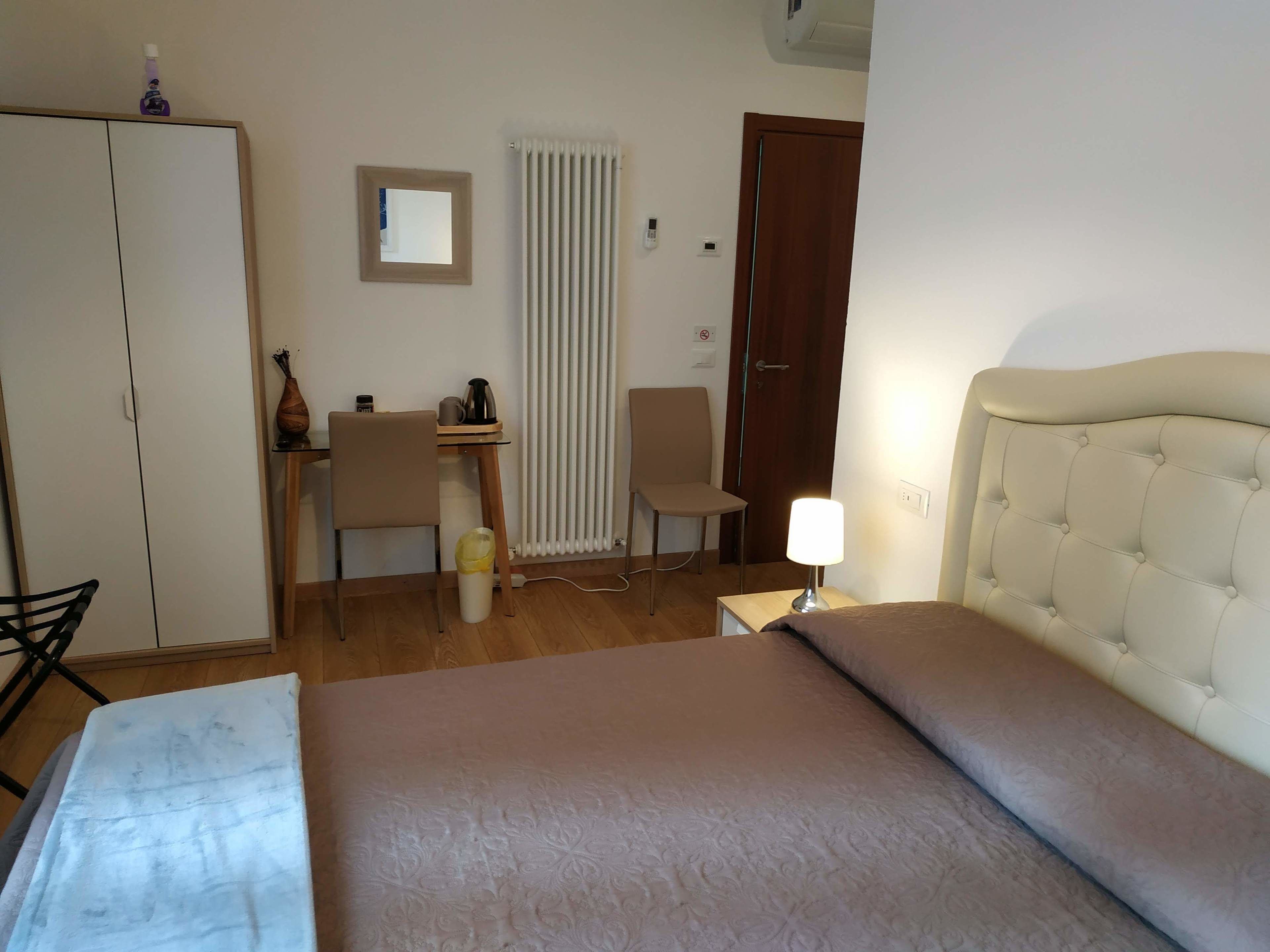 A hotel room with a bed, a desk, a chair, a radiator, and a wardrobe.