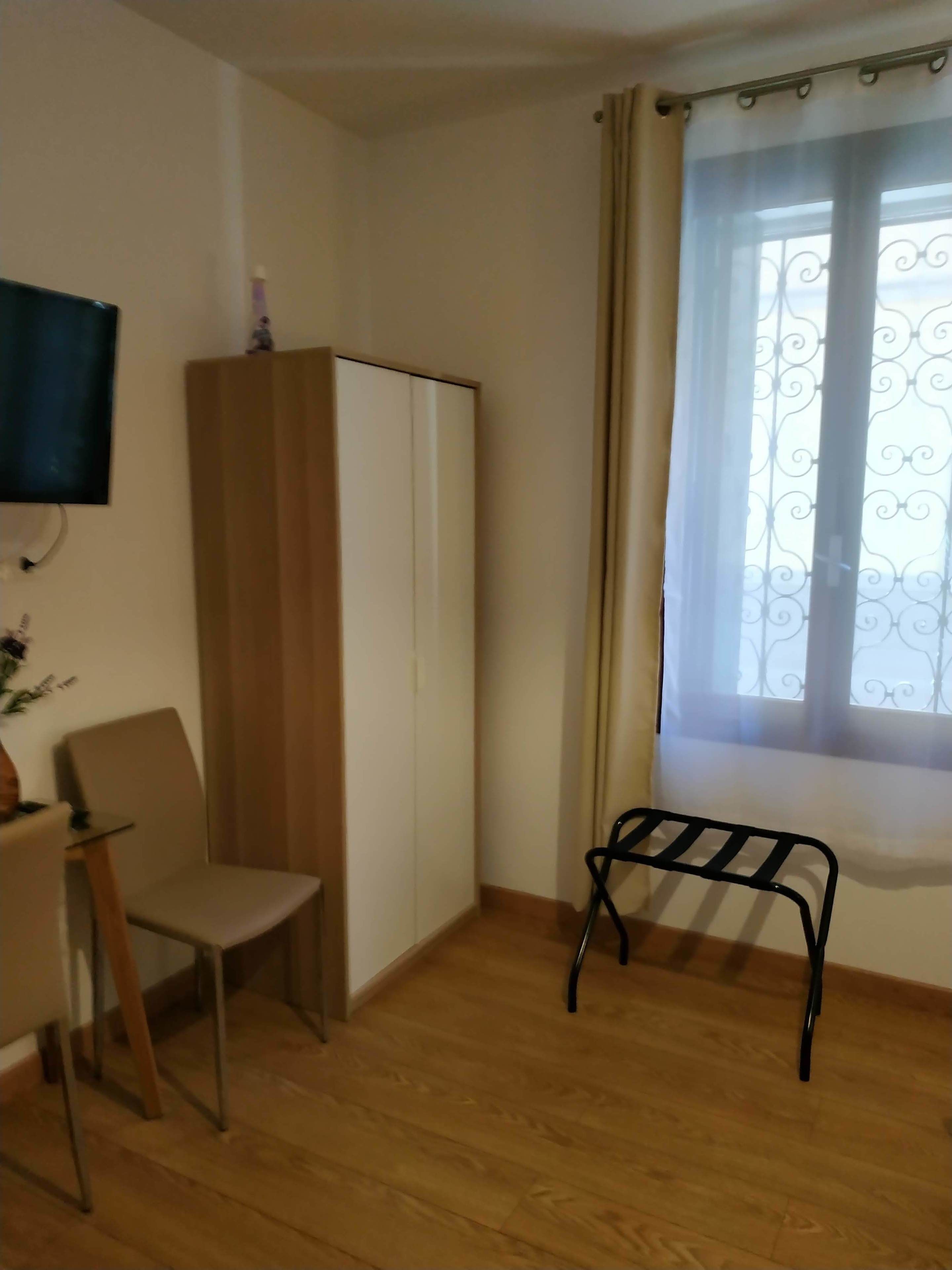 Une pièce avec un plancher en bois, un bureau avec une chaise, une armoire, un téléviseur fixé au mur et une fenêtre avec des rideaux.