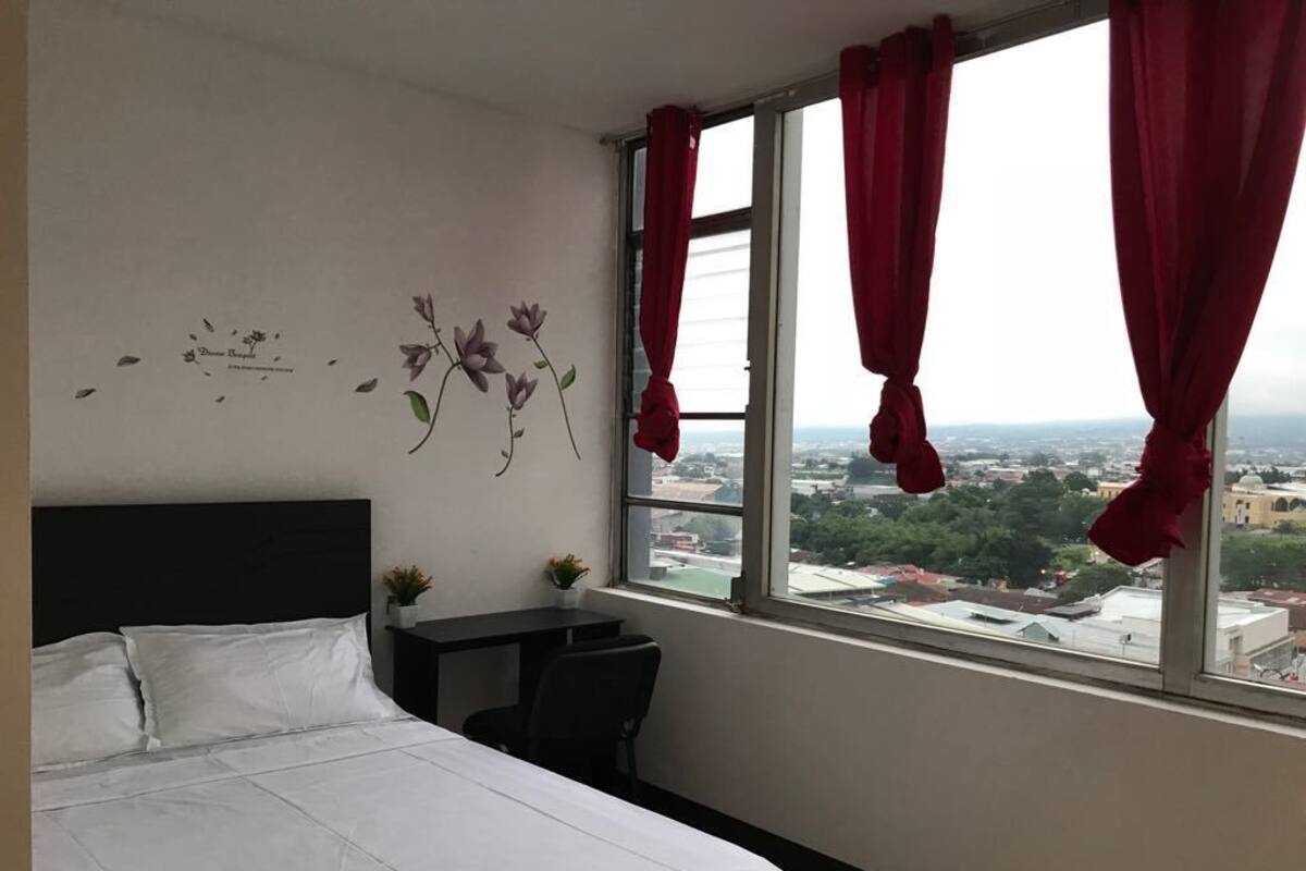 A hotel room with a bed, a desk, and a window with red curtains.
