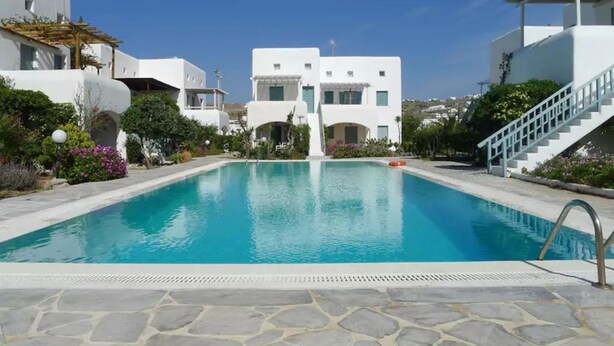 Una piscina con plataforma de piedra, rodeada por edificios blancos con ventanas y balcones arqueados, bajo un cielo azul despejado.