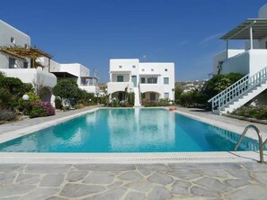 A swimming pool with a stone deck, surrounded by white buildings with arched windows and balconies, under a clear blue sky.