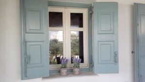 Una ventana con contraventanas abiertas, dos plantas en macetas y vistas al exterior con vegetación.