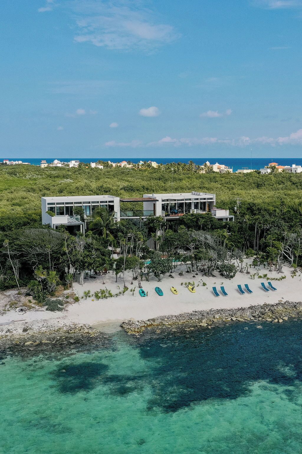 Una propiedad frente a la playa con dos edificios blancos modernos, una playa de arena con kayaks y sillas de descanso, y una vista despejada del océano.