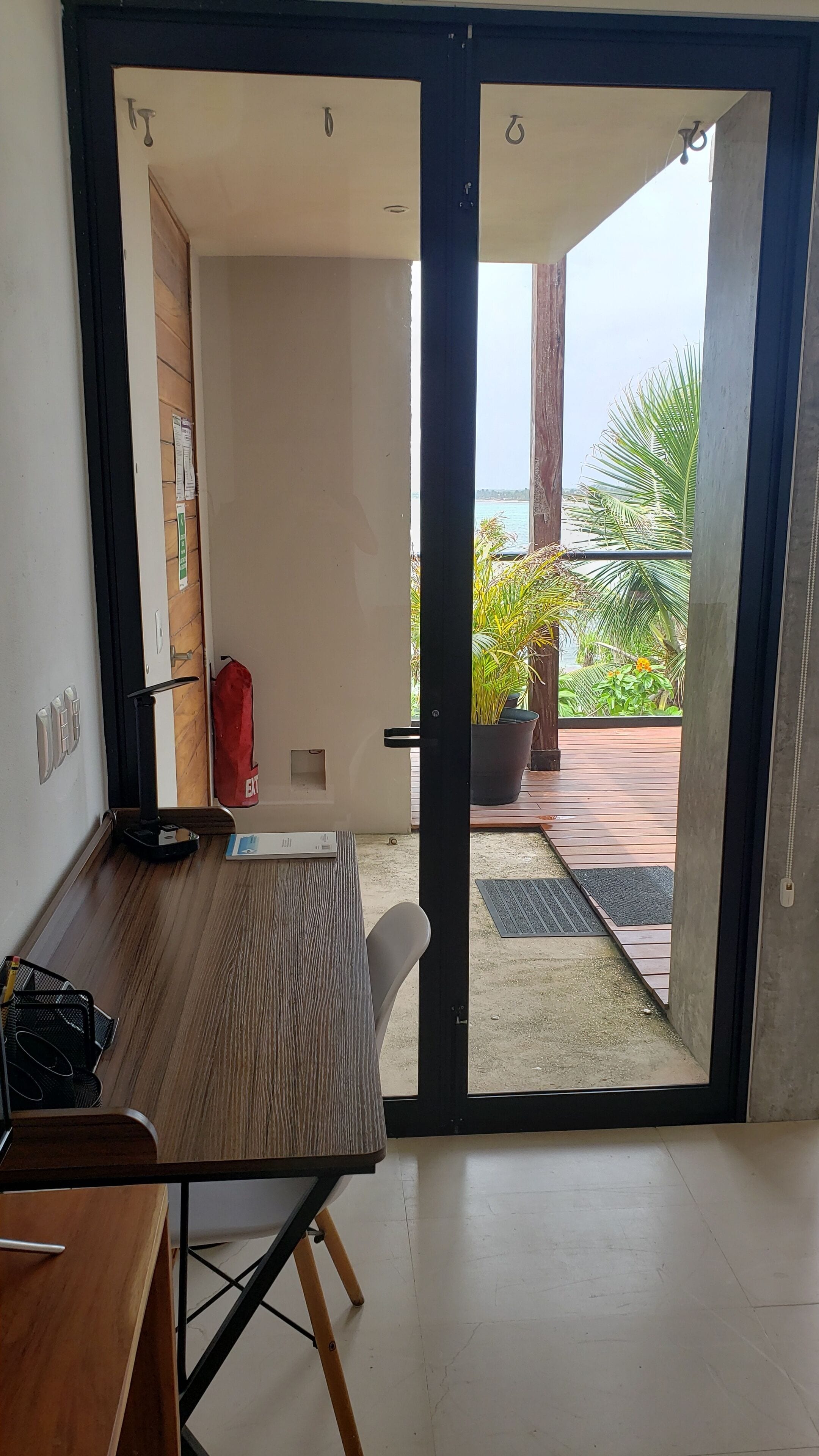 A room with a wooden desk, a chair, a sliding glass door leading to an outdoor area with a wooden deck and potted plants.