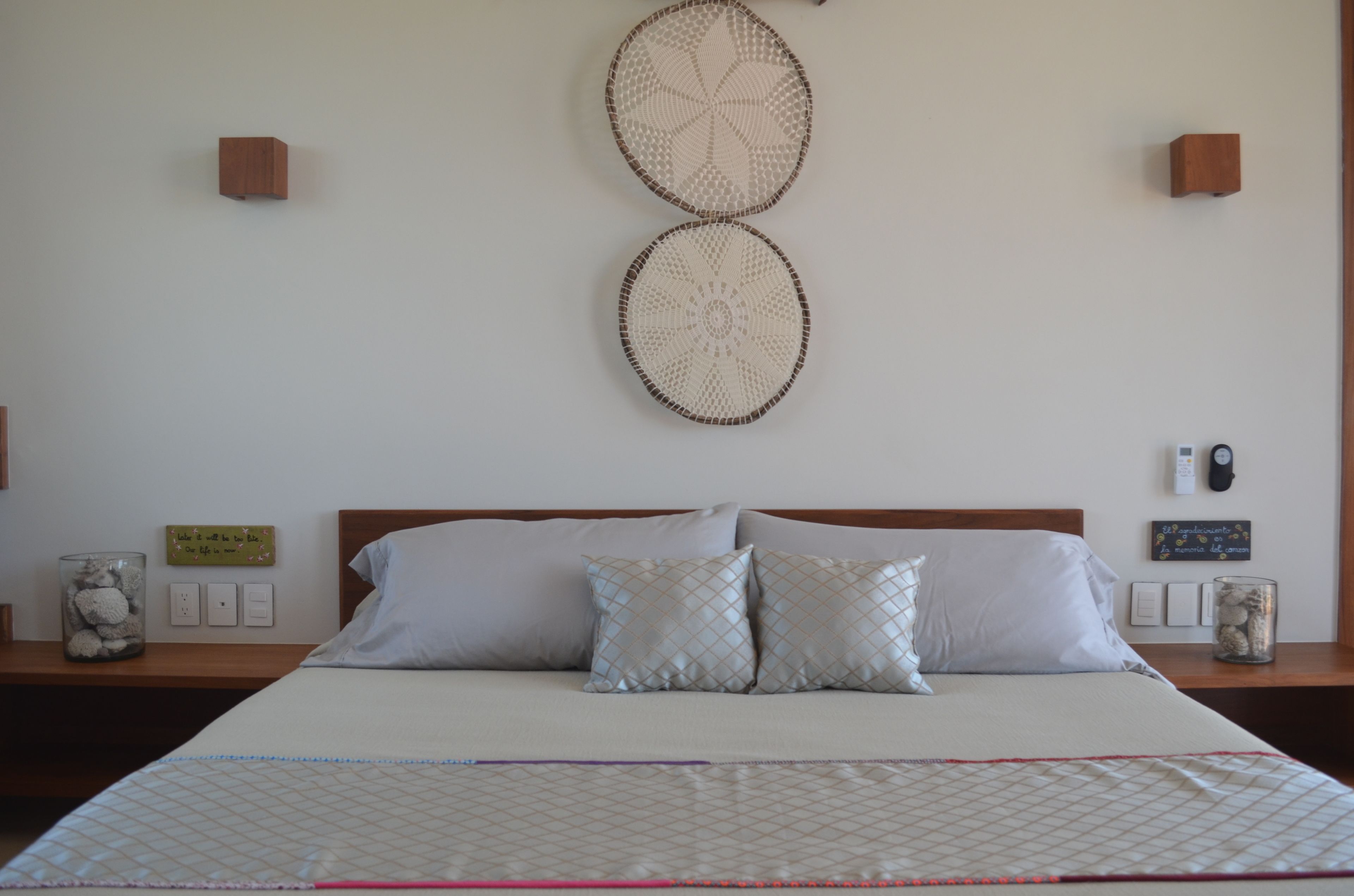 A neatly made bed with a patterned bedspread, two wall-mounted decorative items, bedside tables with glass jars, and a wall clock.