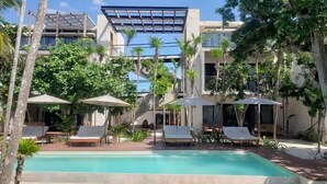 A pool area with lounge chairs and umbrellas, surrounded by palm trees and modern buildings. - Tulsayab Luxury Development (Tulum)