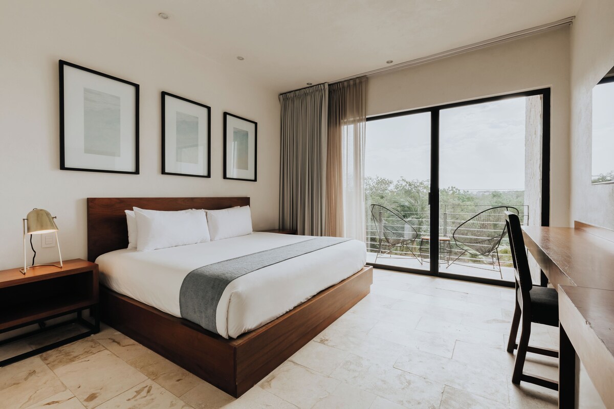 A modern hotel room with a large bed, a desk, and a balcony with a view.