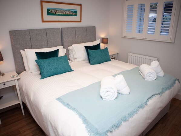 A neatly made bed with white and teal bedding, a framed picture on the wall, and a window with blinds. - 3 Sunday School Court, St Ives - Sleeps 4- Off-Site Parking - Courtyard (St Ives)