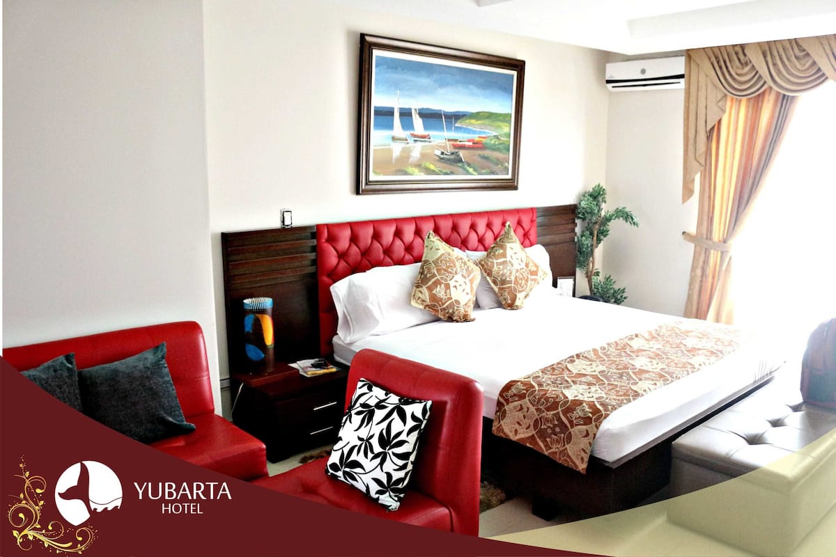 Una habitación de hotel con un sofá tapizado rojo, una cama grande con cabecera roja y un cuadro con una escena costera sobre la cama.
