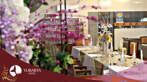 Uma área de refeições com mesas preparadas para o serviço, um bar ao fundo e uma composição floral decorativa.