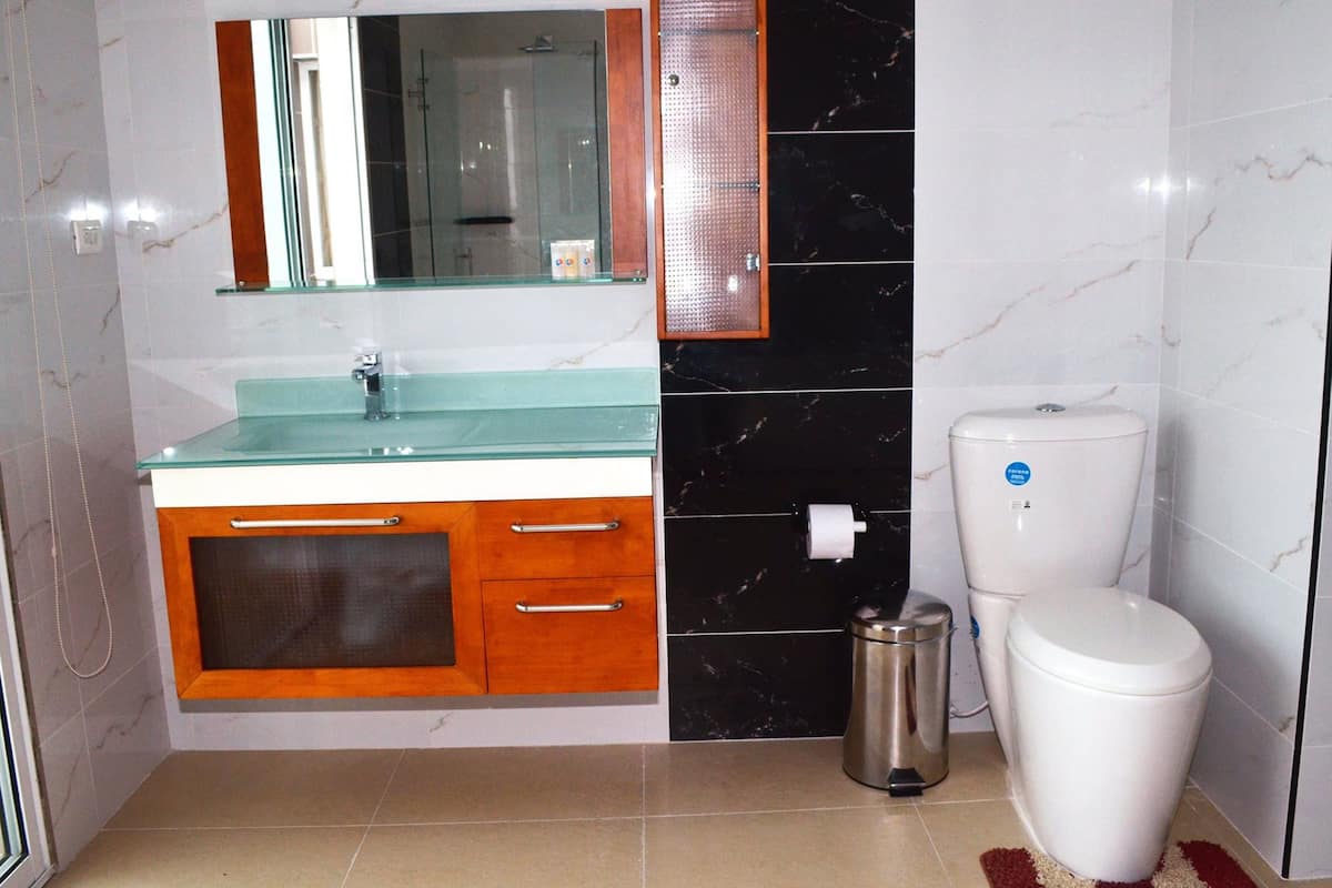 Un baño con lavamanos de vidrio, mueble de madera, baldosas de mármol blanco y negro, inodoro blanco y un cubo de basura.