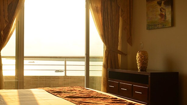 Um quarto com cama, cômoda e vista para o mar através de grandes janelas.