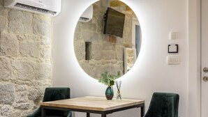 Ein Zimmer mit einer Steinwand, einem runden Spiegel, einem kleinen Tisch mit einer Vase und Räucherstäbchen sowie zwei grünen Stühlen.