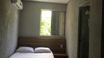 Ein kleiner, schmaler Raum mit einem Einzelbett, einem Fenster mit Jalousien und einer Tür mit einem gemusterten Design.