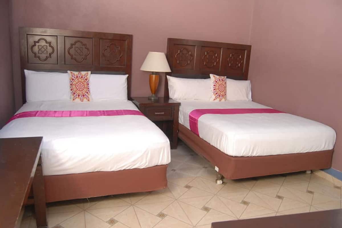 Een hotelkamer met twee bedden, elk met een houten hoofdbord en een roze sprei. Daartussen staat een nachtkastje met een lamp.