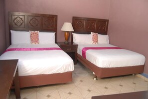 Habitación de hotel con dos camas, cada una con cabecero de madera y colcha rosa. Hay una mesita de noche con lámpara entre las camas.