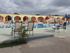 Zona de piscina con sillones, una piscina pequeña y un edificio con ventanas de arco en el fondo.