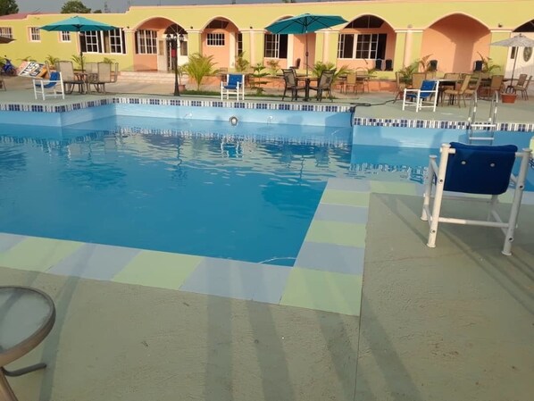 Una piscina con sillas de descanso y sombrillas, rodeada por un edificio con ventanas arqueadas y un suelo de baldosas.