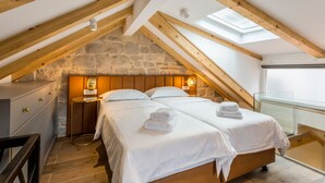 Ein Schlafzimmer mit einer Steinwand, Holzbalken, einem Oberlicht, zwei Betten, Nachttischen und Blick ins Freie.