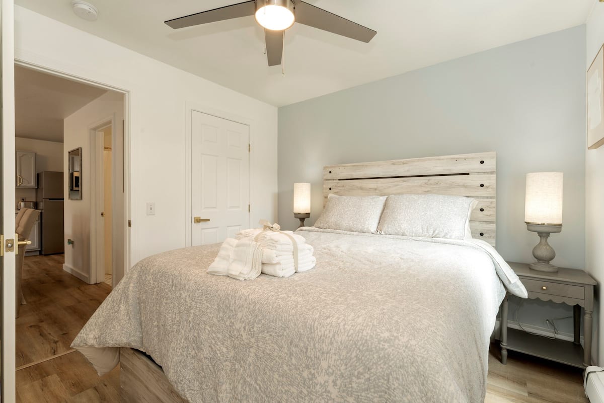 Un dormitorio con cama, ventilador de techo, mesita de noche con lámpara y puerta que da a un baño.