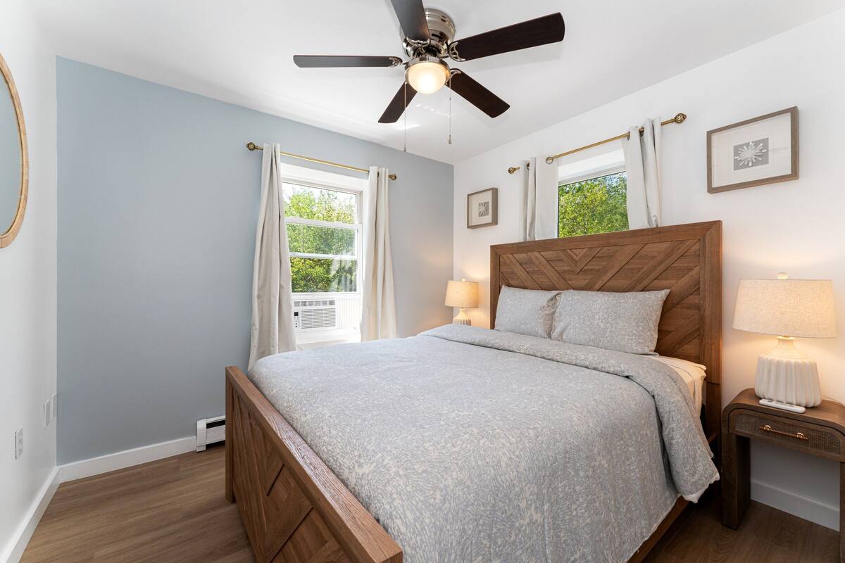 Un dormitorio con cama, ventilador de techo, lámparas de noche y una ventana con cortinas.