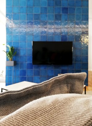 A modern living room with a blue tiled wall, a flat-screen TV mounted above a sofa, and a small plant on the left.