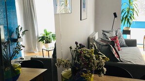 A modern living room with a sofa, a dining table with a vase of flowers, and a large window with a view of the outdoors. - Piper Zajezdnia Blue (Poznan)