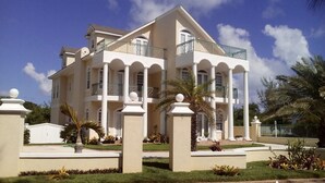 A large, two-story building with a symmetrical facade, multiple balconies, and a well-maintained garden with palm trees and shrubs.