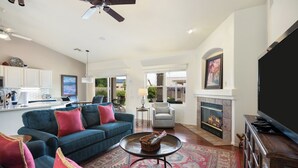A spacious living room with a blue sofa, a fireplace, a television, and a kitchen in the background. - Club Wolff  West - Enjoy Tucson in Style.  Pool, hot tub, outdoor kitchen, view (Tucson)